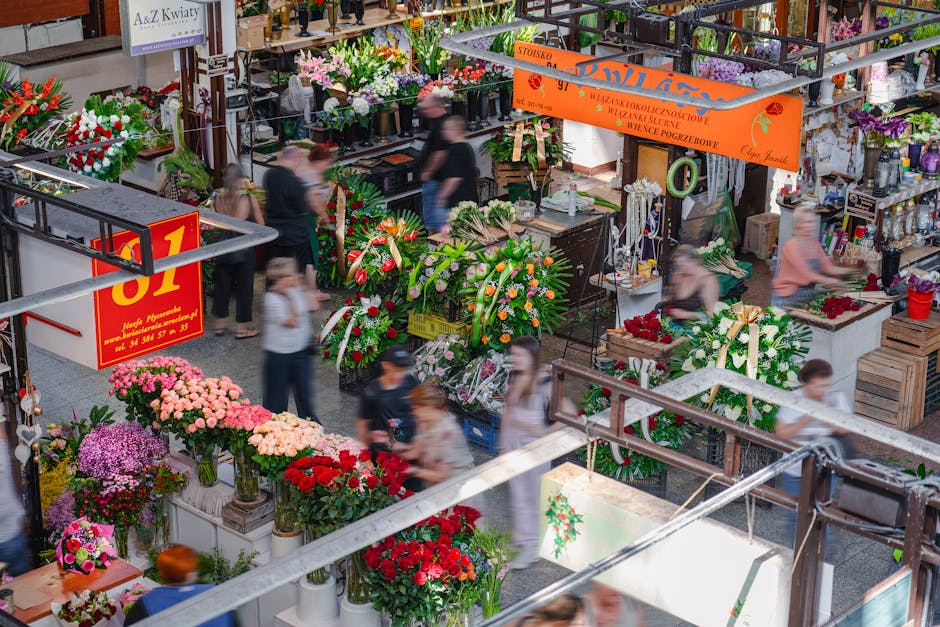 An indoor flower market scene featuring a variety of fresh floral arrangements and bouquets. Prominent in the foreground are vivid pink, red, and purple flowers, including chrysanthemums, roses, and c