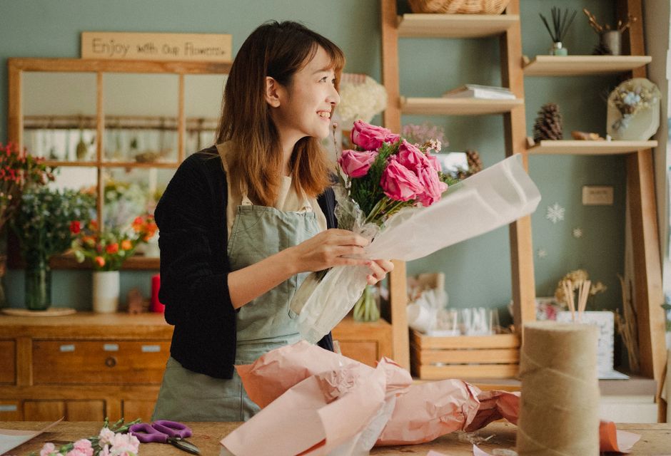 A woman with shoulder-length brown hair, wearing a light gray apron over a black cardigan, is inside a florist shop holding a bouquet of vibrant pink roses and purple tulips wrapped in clear plastic a